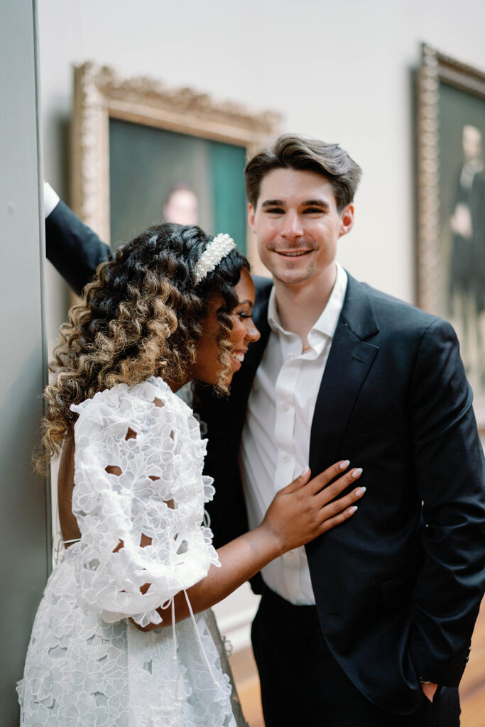Engagement photos of a couple inside the National Gallery of Art Museum in Washington, DC. They sit together on a bench. They are laughing.