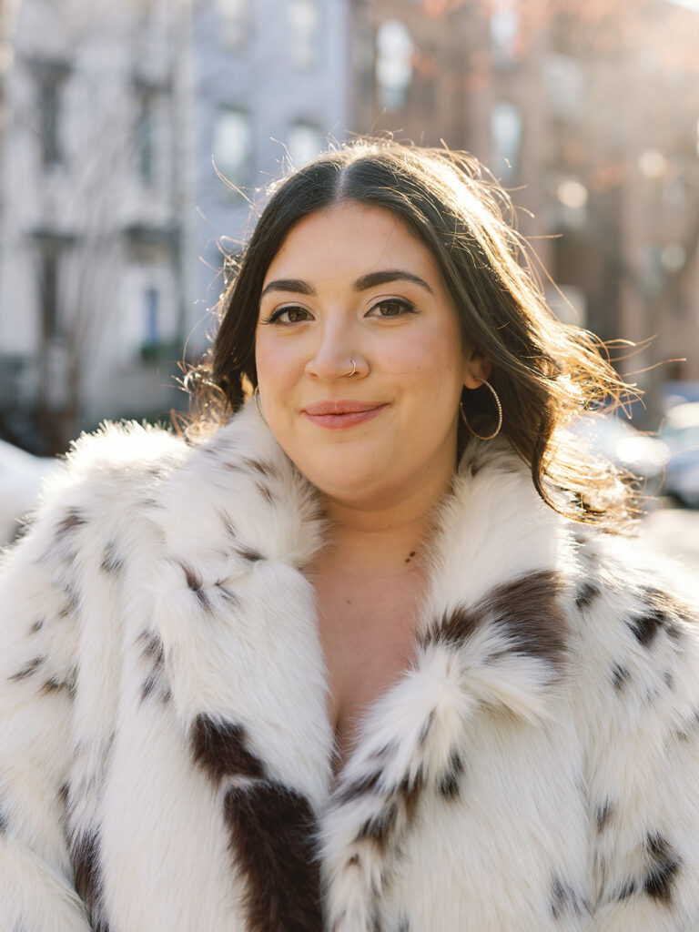 Woman with brown hair and faux fur coat standing in front of brick townhouses for her engagement photos.