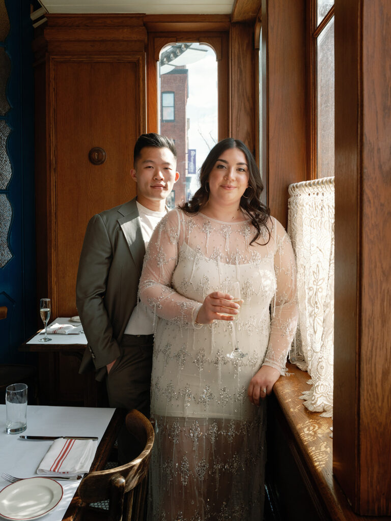 A couple stands together by a window for their engagement photos at Le Diplomate in DC while drinking champagne and holding hands. 