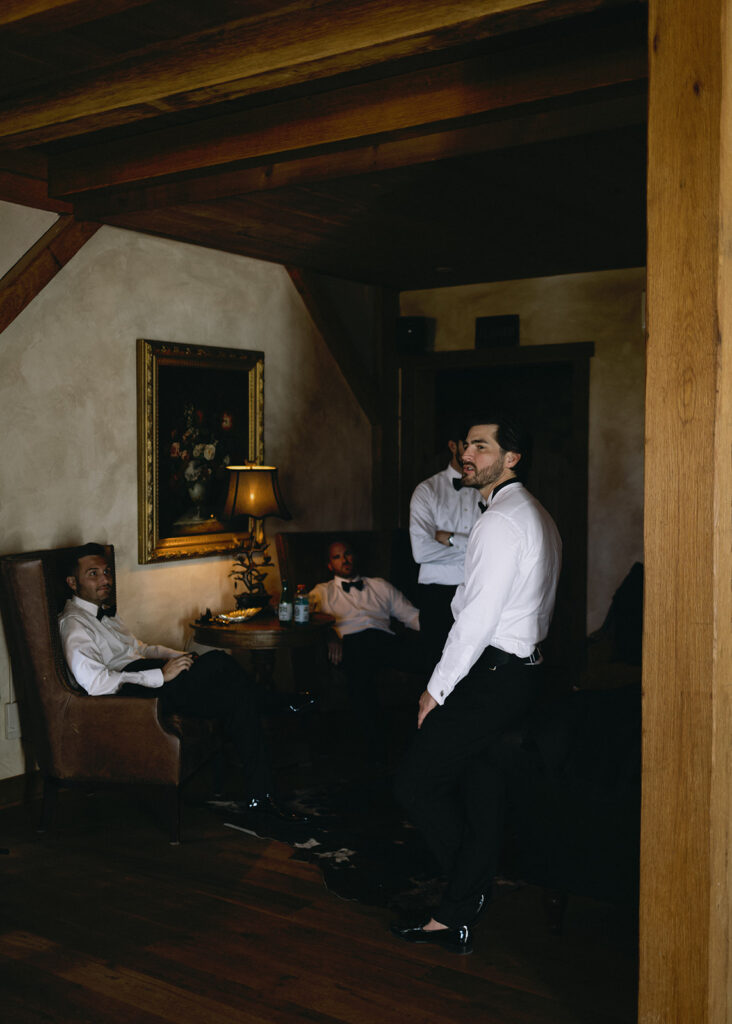 Groomsmen in tuxes inside rustic, moody getting ready room with vintage decor