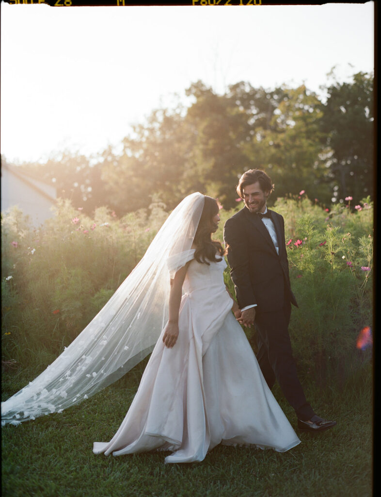 Golden Hour Portrait, Bride and Groom Walking Bride and groom walk hand-in-hand through sunlit wildflowers during golden hour.