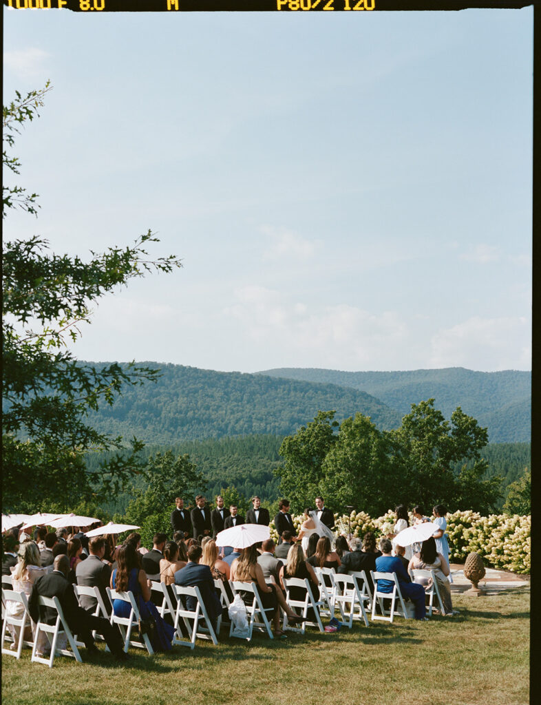 Ceremony Setup with Mountain View Rows of white chairs and a half-moon arrangement of blush flowers overlook a mountain view at an outdoor ceremony.
