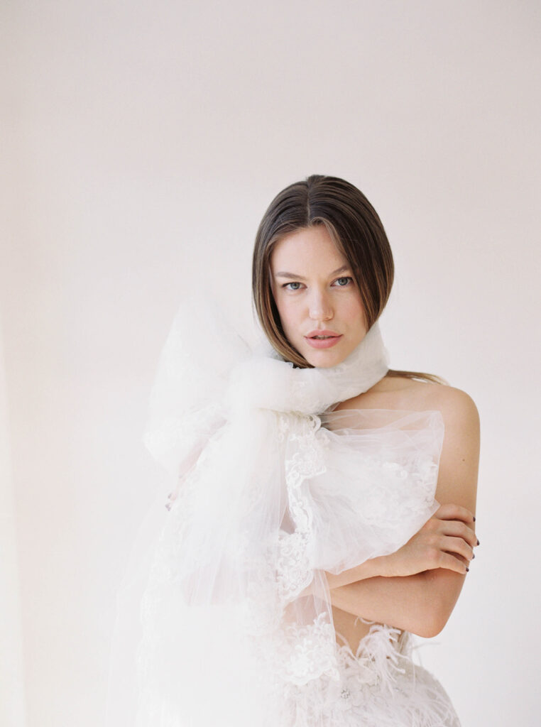 Bride with arms crossed and a feather skirt. She is wearing a wedding veil tied in a bow around her neck. It's large.