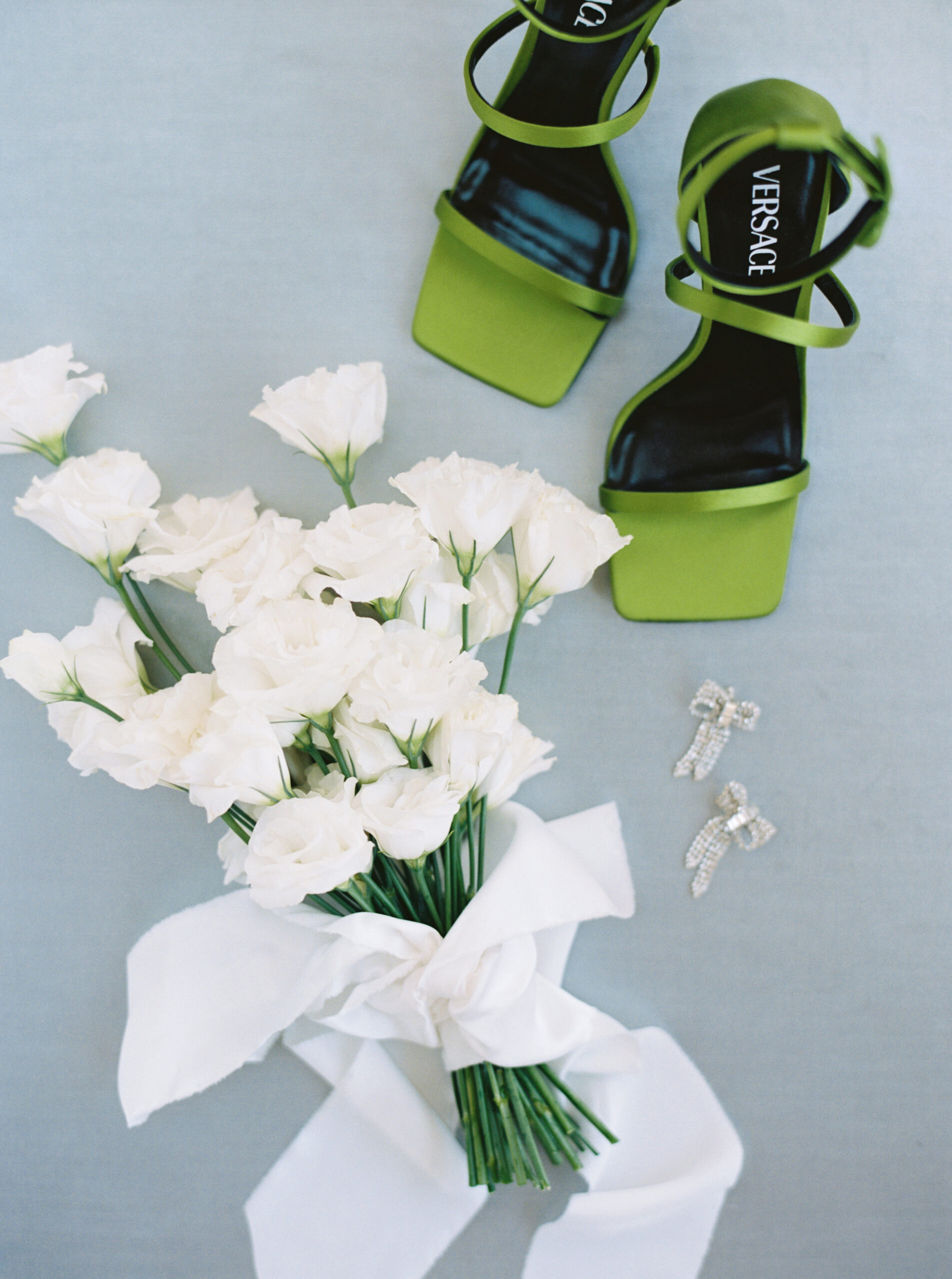 Green versace shoes with a white wedding bouquet and crystal earrings placed on a light blue linen surface