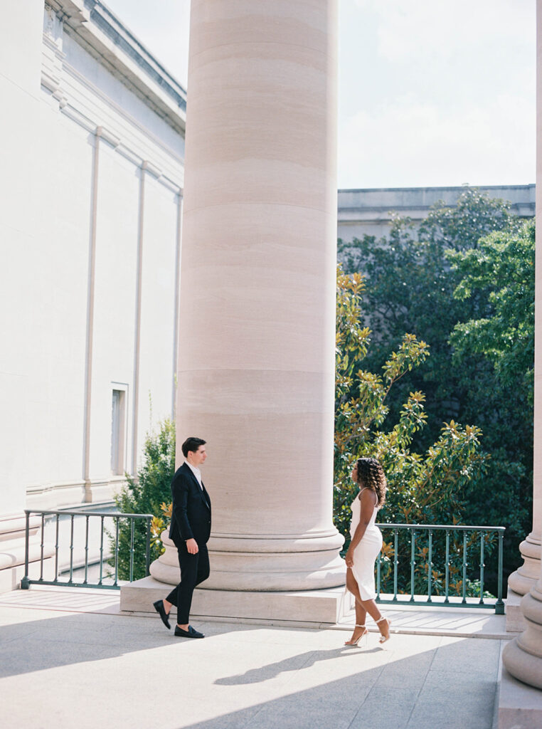 Engagement photos of a couple outside at the National Gallery of Art Museum in Washington, DC. Couple is walking towards one another and their is a building column behind them.