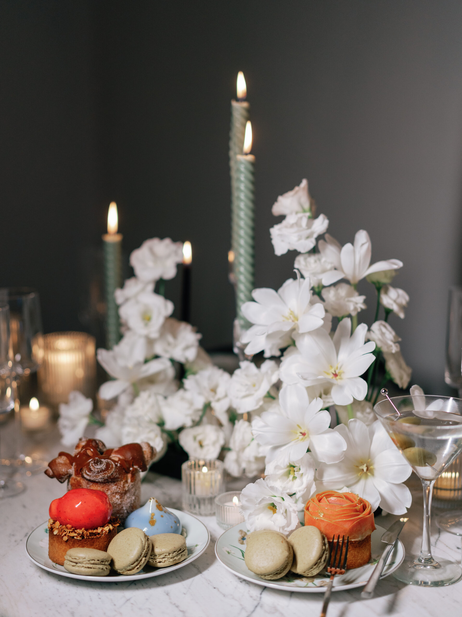 White floral centerpiece with sage green candles on a marble table with pastries and macarons.