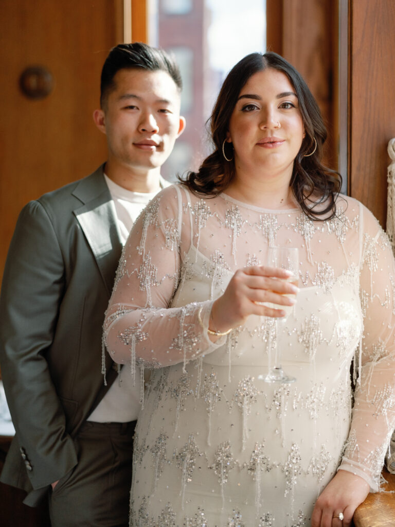 Couple dressed up for engagement photos in front of a wood wall. She is wearing a sparkly dress with a sheer top and he is in a light sage green suit. She is holding a glass of champagne.