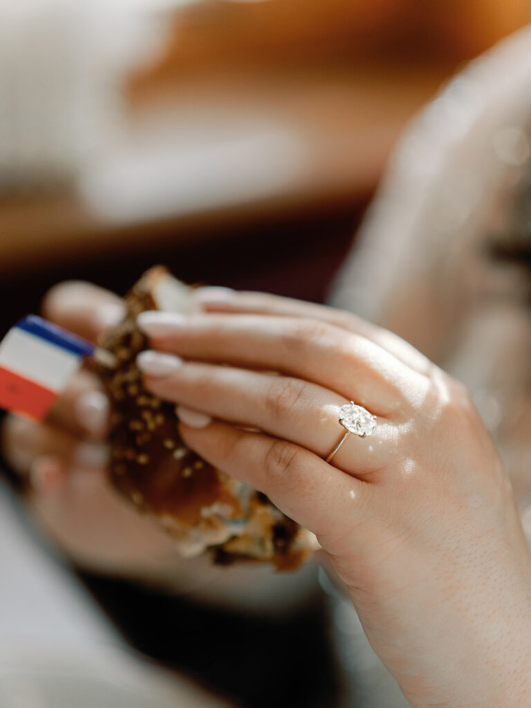 Woman holding a cheeseburger, while the camera is focused on her diamond engagement ring.