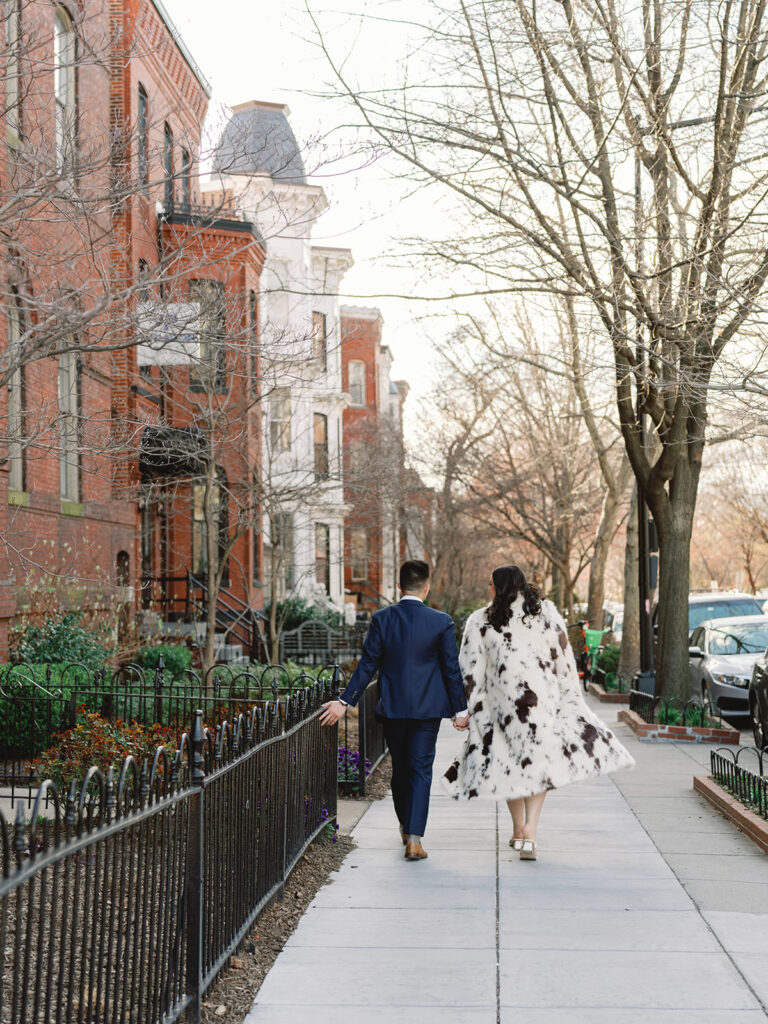 Couple walking down the street holding hands for their engagement photos by brick townhouses. 