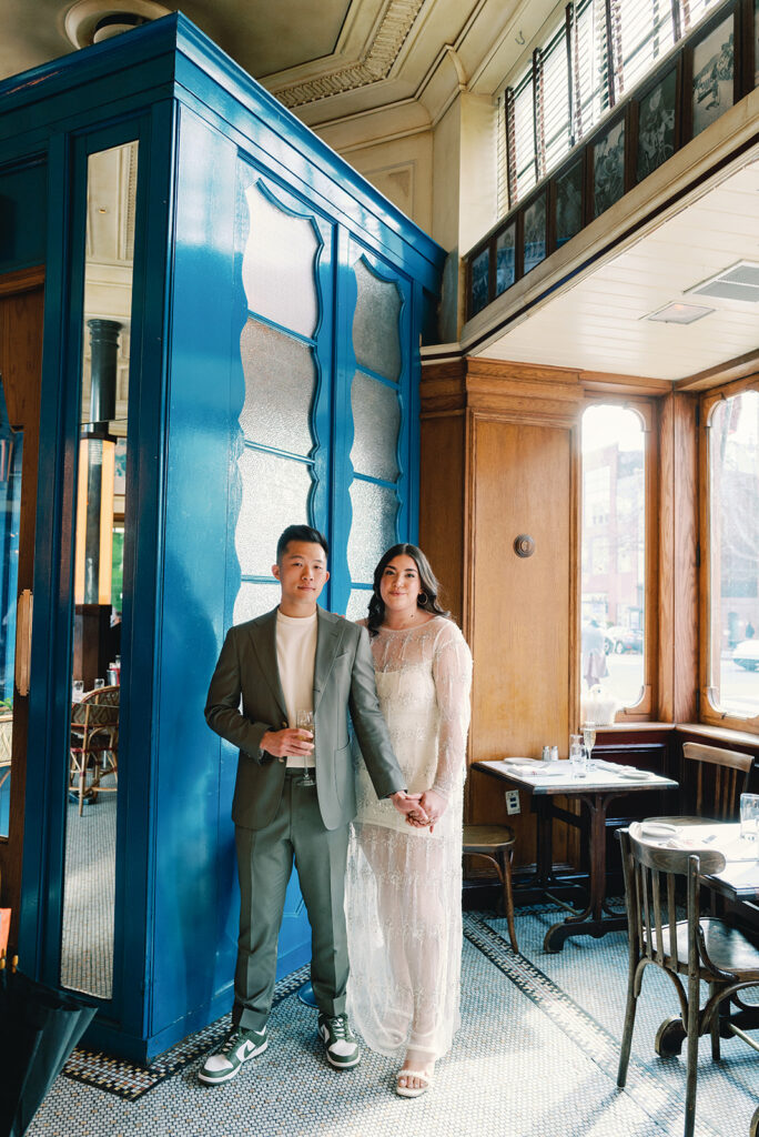 A couple stands together by a window for their engagement photos at Le Diplomate in DC while drinking champagne and holding hands. 