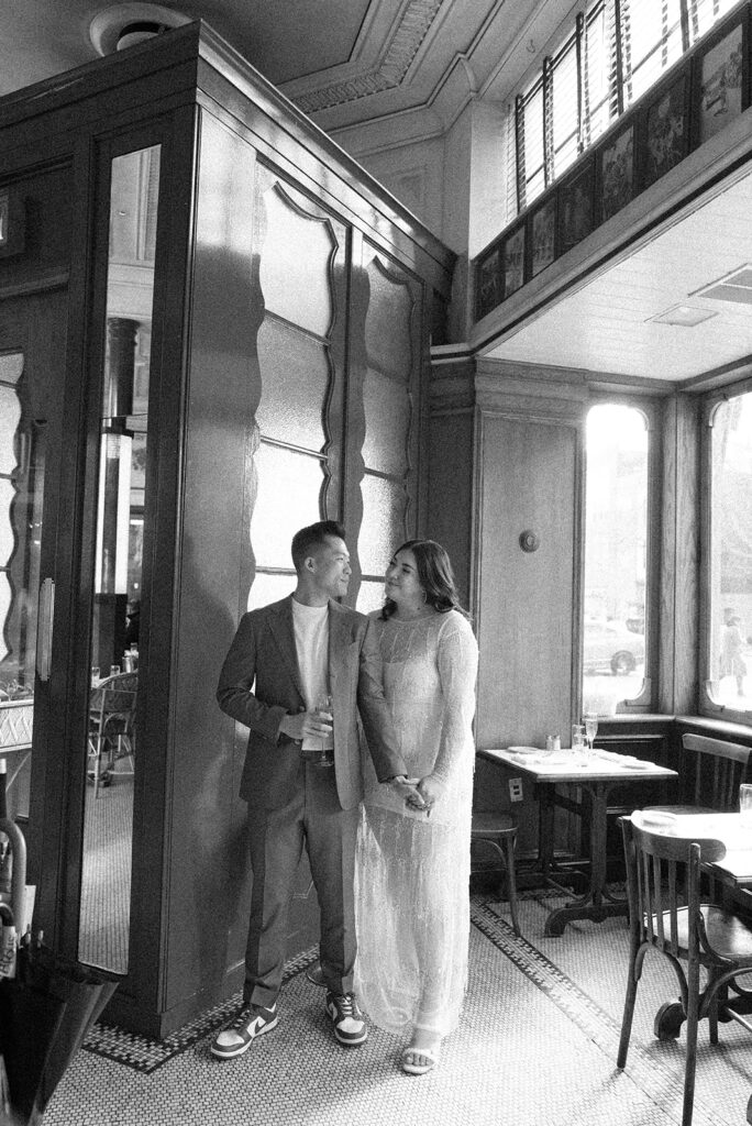 A couple stands together by a window for their engagement photos at Le Diplomate in DC while drinking champagne and holding hands. Black and white photo.