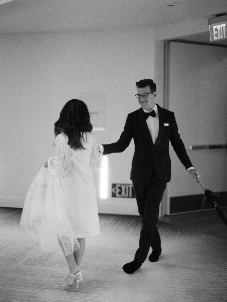 A groom in a classic black tuxedo and bow tie reaches for his bride’s hand as she twirls in a modern, knee-length wedding dress with sheer sleeves. Her delicate veil drapes over her arm as they move together in a softly lit hallway, an 'EXIT' sign glowing in the background. Captured in timeless black and white, this candid moment radiates effortless joy and elegance.