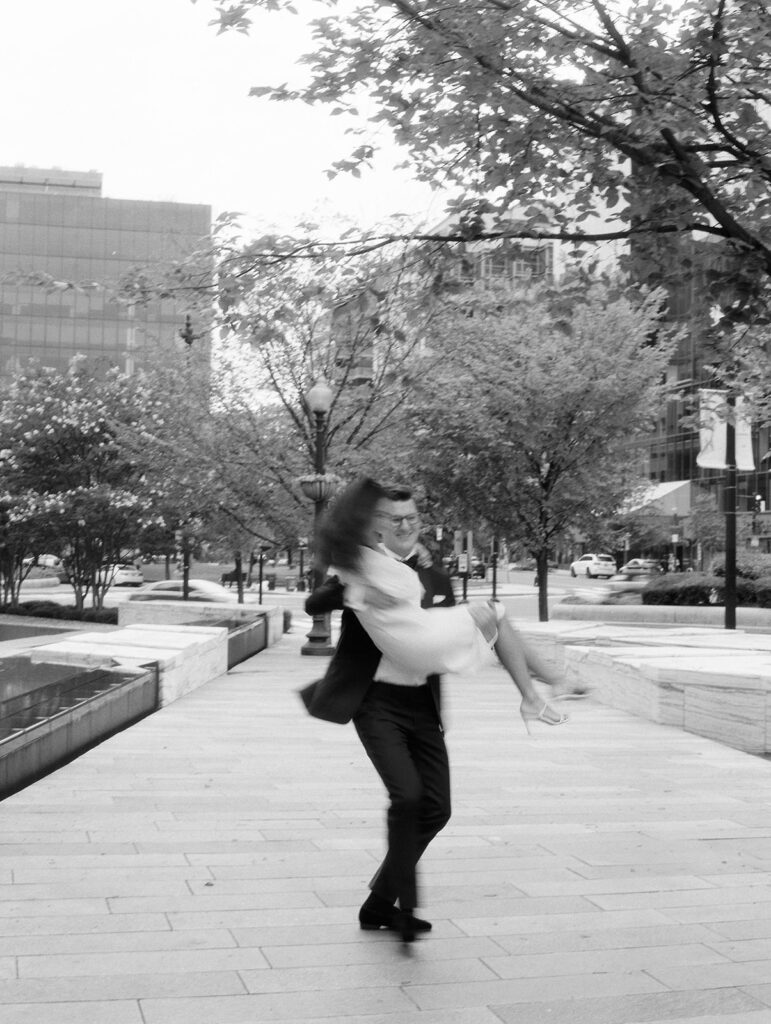 A groom in a sleek black tuxedo twirls his bride in the air on a city sidewalk, her dress billowing as she laughs in motion. Captured in black and white, the couple radiates joy against a backdrop of tree-lined streets, modern glass buildings, and passing cars. A spontaneous, cinematic moment that feels both timeless and full of life.