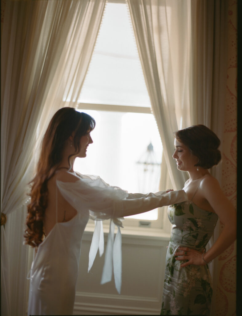 Bridesmaid and bride getting ready on the morning of a wedding in front of a window. The bridesmaid has a tear in her eye and the bride's hands are on the bridesmaid's shoulders. 