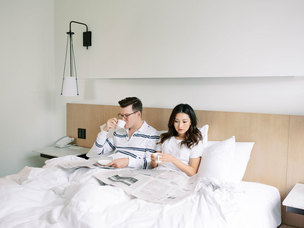 A couple enjoys a quiet morning in bed, wrapped in white linens, sipping espresso while reading the newspaper. The groom, wearing a striped long-sleeve shirt, takes a sip from his cup, while the bride, in a simple white tee, gazes at her drink with a serene expression. The minimalist, modern bedroom setting with soft natural light evokes an effortless, intimate start to their wedding day.