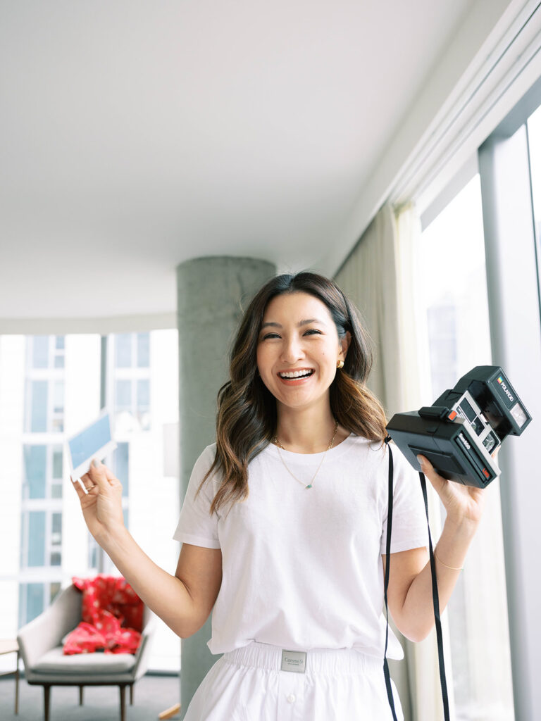 A bride beams with joy, holding a vintage Polaroid camera in one hand and a freshly printed instant photo in the other. Dressed in a casual white tee and soft cotton lounge pants, she stands near a bright floor-to-ceiling window, bathed in natural light. The playful, carefree energy of the morning sets the tone for an effortlessly stylish wedding day.