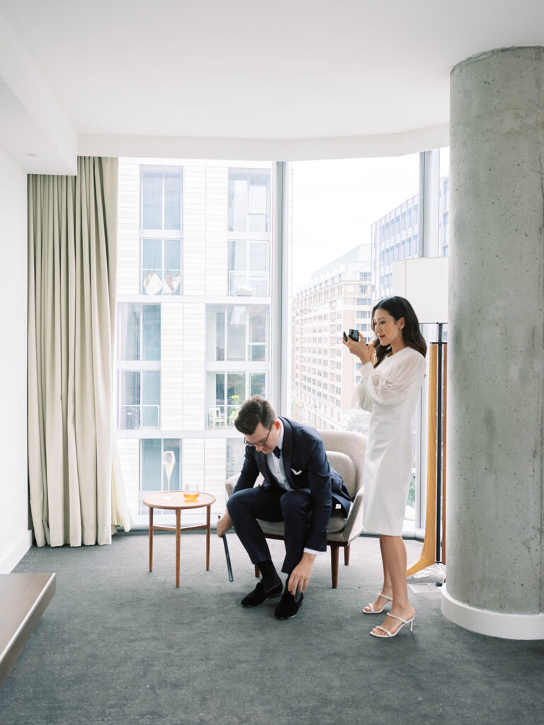 A groom in a navy tuxedo and black loafers sits in a modern hotel room, using a shoehorn to slip on his shoes. His hair is neatly styled, and he wears glasses. Behind him, a woman in a white, knee-length dress with sheer, billowing sleeves stands with a small camera, capturing the moment. She wears delicate white heels. The room features floor-to-ceiling windows with views of contemporary city buildings, a concrete column, and a minimalist interior design. A small round table nearby holds a champagne flute and a glass of amber-colored liquid.