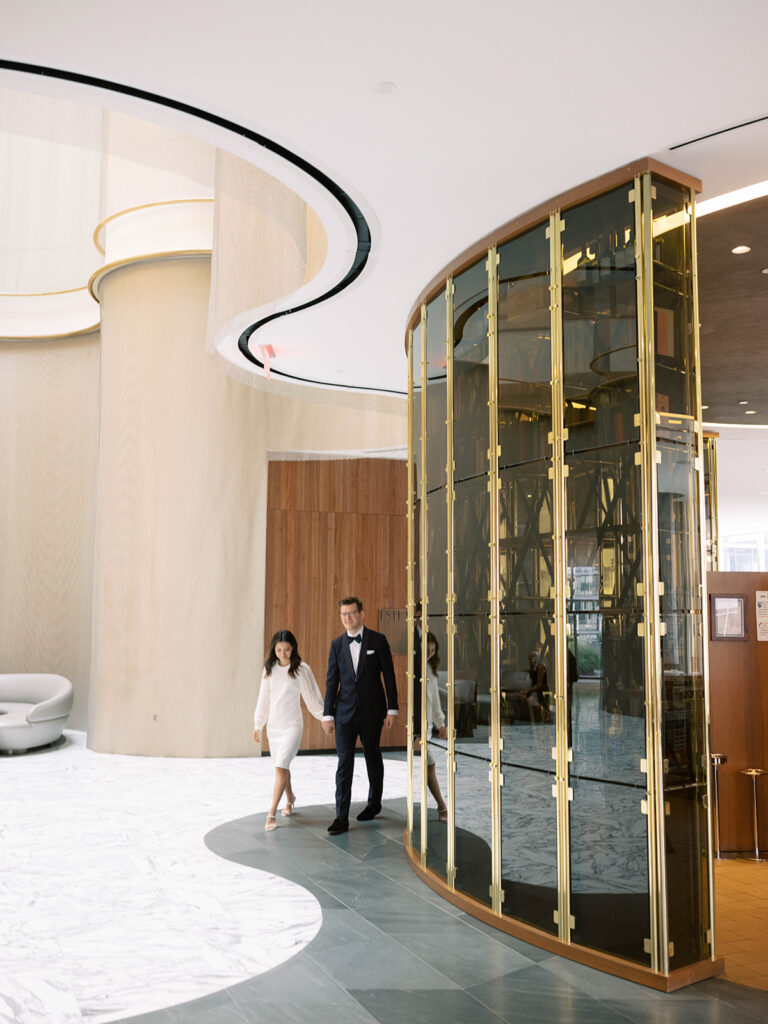 A bride and groom walk hand in hand through the sleek, modern lobby of The Conrad Hotel in Washington, DC. The bride wears a chic white dress with sheer billowing sleeves, while the groom looks sharp in a classic black tuxedo. The grand curved architecture, marble floors, and golden glass wine display create a luxurious, contemporary backdrop for their effortlessly elegant wedding day.